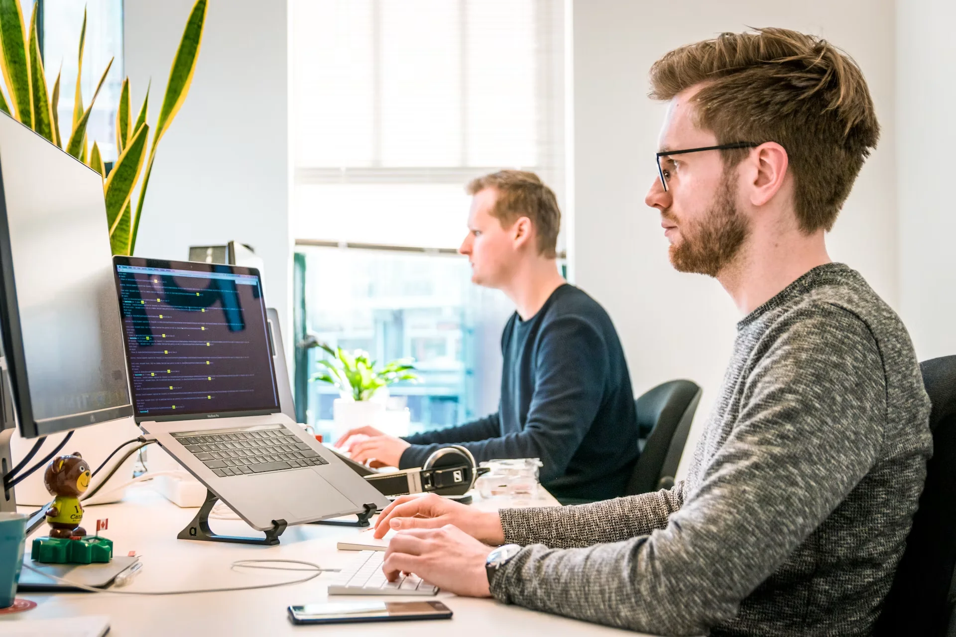 Dois homens em um escritório, trabalhando em computadores. Um deles digita no teclado, o outro está ao fundo